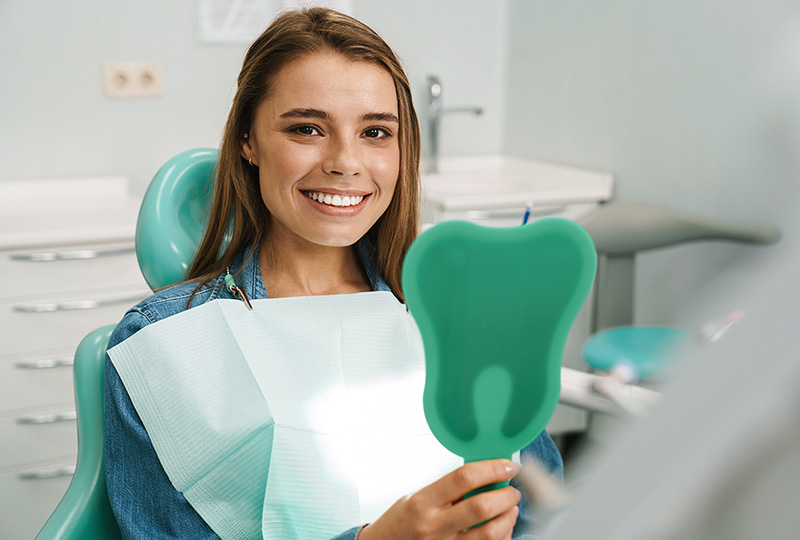 A young woman is smiling at the camera while seated in a dental chair, holding a green object with her right hand, which appears to be an oral hygiene tool or device, possibly for cleaning teeth or measuring mouth size. She is wearing a light blue denim jacket and has her hair tied back. The setting suggests she is in a dental office, as indicated by the presence of dental equipment and the professional attire of the person holding the object.