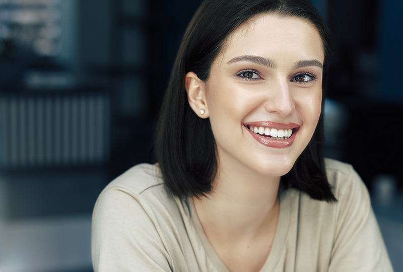 The image features a woman with short hair smiling at the camera, wearing a light-colored top, with a neutral background that includes what appears to be a shelf or display area.