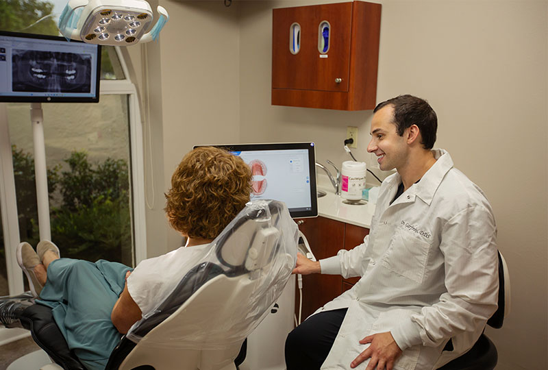 The image shows a man sitting in a dental chair, holding up a mirror to his face, with a beard and mustache, wearing a green vest, presumably a dentist or dental hygienist.