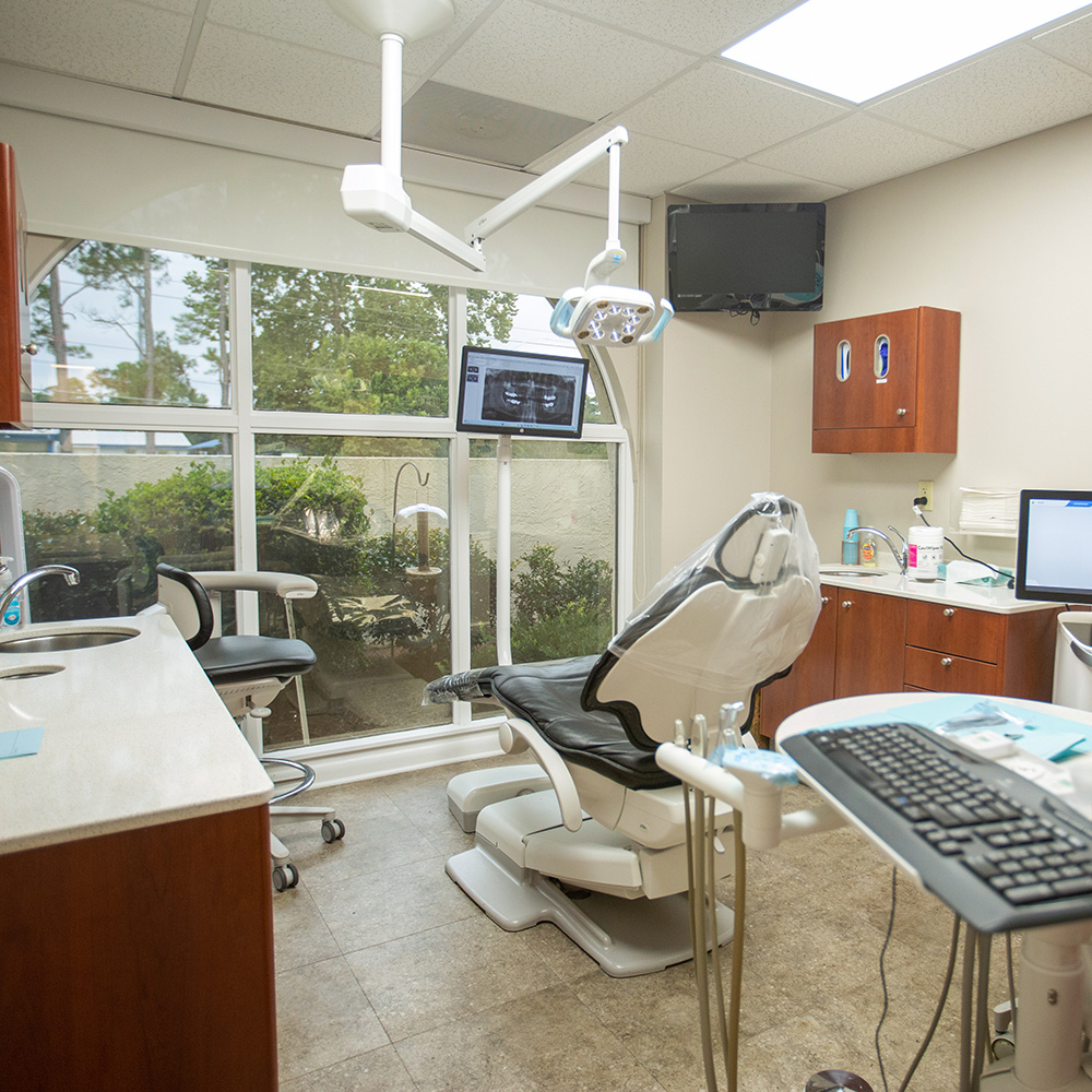 This image shows a person sitting in a dental chair, smiling at the camera while wearing a blue surgical mask, with their mouth open as if they are receiving dental care, and there s a dental hygiene tool visible near them.